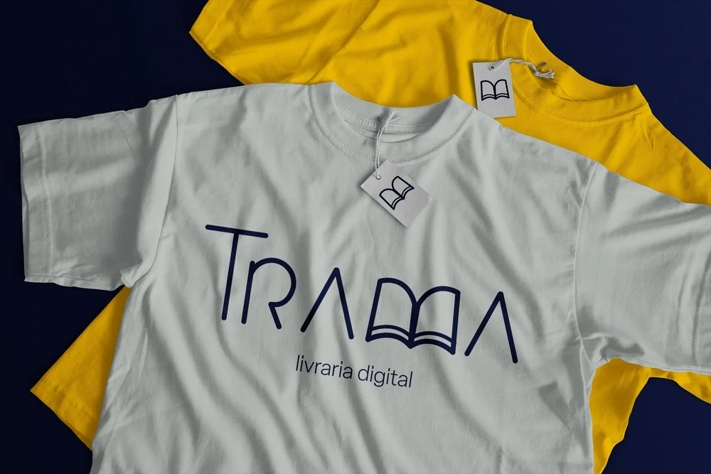 Duas camisetas, uma cinza-clara e outra amarela, com o logo da Trama e uma etiqueta com o símbolo do livro aberto